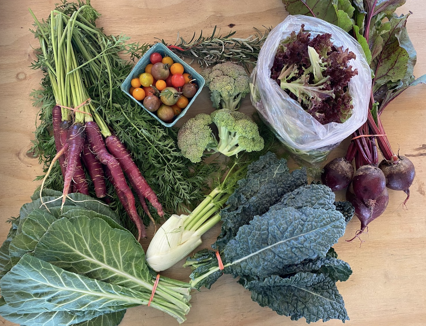 Alicia - Screen Shot 2023-06-06 at 5.43.36 PM Fresh assortment of organic vegetables including purple carrots, broccoli, collard greens, kale, beets, and a variety of cherry tomatoes, displayed on a wooden surface, highlighting the benefits of healthy eating and local produce.